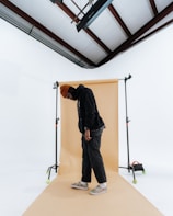 A person is standing in a photography studio setting, with a beige backdrop and a white surrounding area. The person is wearing a dark outfit with a brown beanie and colorful sneakers. There is studio lighting equipment visible, as well as a ceiling with wooden beams.