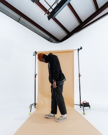 A person is standing in a photography studio setting, with a beige backdrop and a white surrounding area. The person is wearing a dark outfit with a brown beanie and colorful sneakers. There is studio lighting equipment visible, as well as a ceiling with wooden beams.