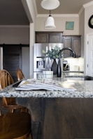 A modern kitchen interior featuring a granite countertop island with an open magazine. Wooden chairs are positioned nearby. Stainless steel appliances, including a refrigerator and a sleek black faucet, add a contemporary touch. A vase with green leaves sits on the island, and there is subtle decor on the walls, including a clock and a small painting. The lighting creates a warm and welcoming atmosphere.