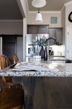 A modern kitchen interior featuring a granite countertop island with an open magazine. Wooden chairs are positioned nearby. Stainless steel appliances, including a refrigerator and a sleek black faucet, add a contemporary touch. A vase with green leaves sits on the island, and there is subtle decor on the walls, including a clock and a small painting. The lighting creates a warm and welcoming atmosphere.