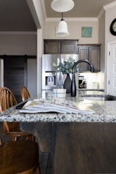 A modern kitchen interior featuring a granite countertop island with an open magazine. Wooden chairs are positioned nearby. Stainless steel appliances, including a refrigerator and a sleek black faucet, add a contemporary touch. A vase with green leaves sits on the island, and there is subtle decor on the walls, including a clock and a small painting. The lighting creates a warm and welcoming atmosphere.