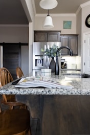A modern kitchen interior featuring a granite countertop island with an open magazine. Wooden chairs are positioned nearby. Stainless steel appliances, including a refrigerator and a sleek black faucet, add a contemporary touch. A vase with green leaves sits on the island, and there is subtle decor on the walls, including a clock and a small painting. The lighting creates a warm and welcoming atmosphere.