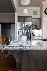 A modern kitchen interior featuring a granite countertop island with an open magazine. Wooden chairs are positioned nearby. Stainless steel appliances, including a refrigerator and a sleek black faucet, add a contemporary touch. A vase with green leaves sits on the island, and there is subtle decor on the walls, including a clock and a small painting. The lighting creates a warm and welcoming atmosphere.