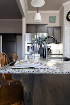 A modern kitchen interior featuring a granite countertop island with an open magazine. Wooden chairs are positioned nearby. Stainless steel appliances, including a refrigerator and a sleek black faucet, add a contemporary touch. A vase with green leaves sits on the island, and there is subtle decor on the walls, including a clock and a small painting. The lighting creates a warm and welcoming atmosphere.