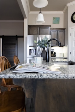 A modern kitchen interior featuring a granite countertop island with an open magazine. Wooden chairs are positioned nearby. Stainless steel appliances, including a refrigerator and a sleek black faucet, add a contemporary touch. A vase with green leaves sits on the island, and there is subtle decor on the walls, including a clock and a small painting. The lighting creates a warm and welcoming atmosphere.