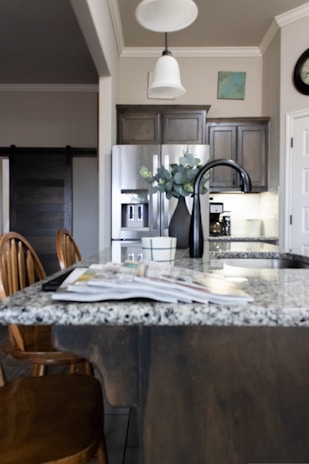 A modern kitchen interior featuring a granite countertop island with an open magazine. Wooden chairs are positioned nearby. Stainless steel appliances, including a refrigerator and a sleek black faucet, add a contemporary touch. A vase with green leaves sits on the island, and there is subtle decor on the walls, including a clock and a small painting. The lighting creates a warm and welcoming atmosphere.