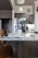 A modern kitchen interior featuring a granite countertop island with an open magazine. Wooden chairs are positioned nearby. Stainless steel appliances, including a refrigerator and a sleek black faucet, add a contemporary touch. A vase with green leaves sits on the island, and there is subtle decor on the walls, including a clock and a small painting. The lighting creates a warm and welcoming atmosphere.