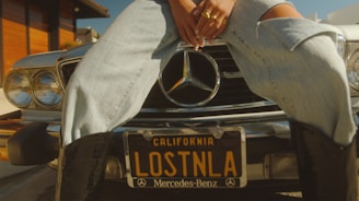 A person sits on the front bumper of a vintage Mercedes-Benz car with a California license plate reading 'LOSTNLA.' The individual is wearing ripped jeans and rings, and the setting conveys a sunny, urban environment.