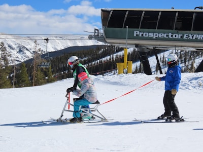 Two skiers on a snow-covered slope, one sitting on a ski chair and the other standing, holding a tether. Both are wearing helmets and winter gear. A ski lift is visible in the background, along with snow-covered trees and mountains.