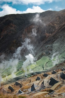 A mountainous landscape with steam rising from various parts of the terrain, indicative of geothermal activity. The ground is a mix of earthy browns and greens, with white steam creating a contrasting element. There are marked pathways and structures that suggest a managed or tourist-friendly area.