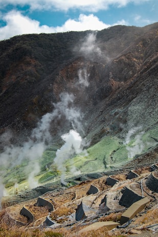 A mountainous landscape with steam rising from various parts of the terrain, indicative of geothermal activity. The ground is a mix of earthy browns and greens, with white steam creating a contrasting element. There are marked pathways and structures that suggest a managed or tourist-friendly area.
