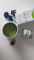 A cup filled with green liquid, likely a matcha beverage, is placed on a white surface. Nearby, there is a tin labeled 'matcha' and a carton of organic soy beverage. A bamboo whisk is beside the cup. Three origami paper figures are positioned in the background.