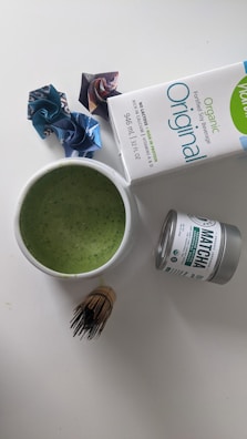 A cup filled with green liquid, likely a matcha beverage, is placed on a white surface. Nearby, there is a tin labeled 'matcha' and a carton of organic soy beverage. A bamboo whisk is beside the cup. Three origami paper figures are positioned in the background.