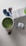 A cup filled with green liquid, likely a matcha beverage, is placed on a white surface. Nearby, there is a tin labeled 'matcha' and a carton of organic soy beverage. A bamboo whisk is beside the cup. Three origami paper figures are positioned in the background.