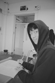 A person wearing a hooded sweatshirt sits at a desk writing with a pen on a piece of paper. The room has a minimalistic design with a closed door in the background and a waste basket on the floor nearby. The overall ambiance is casual and informal.