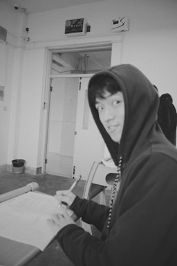 A person wearing a hooded sweatshirt sits at a desk writing with a pen on a piece of paper. The room has a minimalistic design with a closed door in the background and a waste basket on the floor nearby. The overall ambiance is casual and informal.