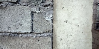 A close-up of a wall section showing a contrast between rough, textured concrete blocks on the left and a smoother plastered surface on the right. The divide is marked by visible cracks and weathering.