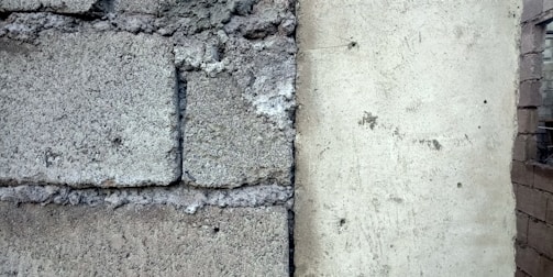A close-up of a wall section showing a contrast between rough, textured concrete blocks on the left and a smoother plastered surface on the right. The divide is marked by visible cracks and weathering.