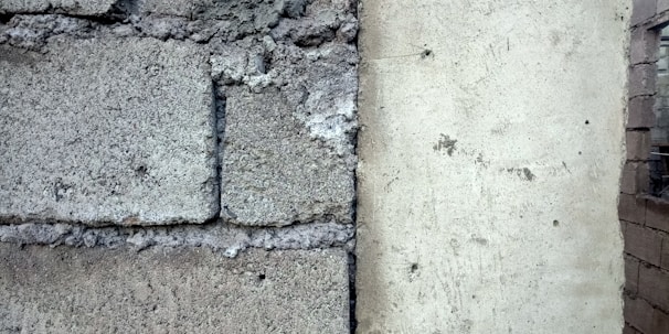 A close-up of a wall section showing a contrast between rough, textured concrete blocks on the left and a smoother plastered surface on the right. The divide is marked by visible cracks and weathering.
