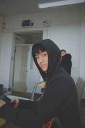 A person wearing a black hoodie sits at a desk in a classroom setting, holding a tablet device. The room has white walls and a fluorescent light fixture on the ceiling. Another person in a black hoodie is in the background, facing away.