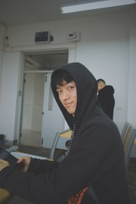 A person wearing a black hoodie sits at a desk in a classroom setting, holding a tablet device. The room has white walls and a fluorescent light fixture on the ceiling. Another person in a black hoodie is in the background, facing away.