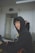 A person wearing a black hoodie sits at a desk in a classroom setting, holding a tablet device. The room has white walls and a fluorescent light fixture on the ceiling. Another person in a black hoodie is in the background, facing away.