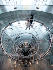 A modern, transparent spiral staircase constructed of glass and metal, viewed from above. The staircase creates an intricate pattern with circular and reflective elements. Two people are walking up the stairs, surrounded by the sleek design.