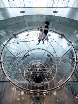 A modern, transparent spiral staircase constructed of glass and metal, viewed from above. The staircase creates an intricate pattern with circular and reflective elements. Two people are walking up the stairs, surrounded by the sleek design.