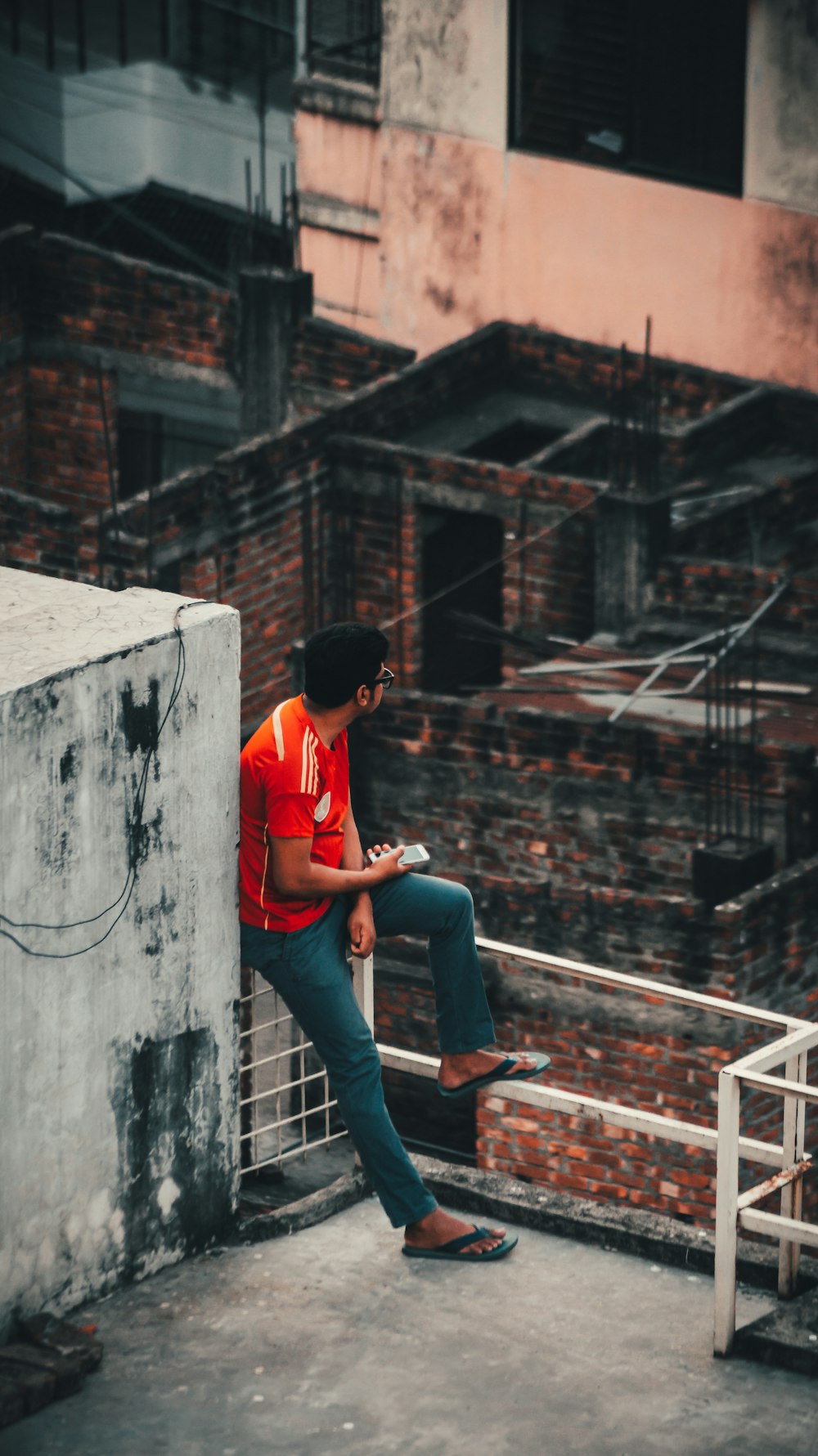 man in red t-shirt and blue denim jeans sitting on concrete wall during daytime