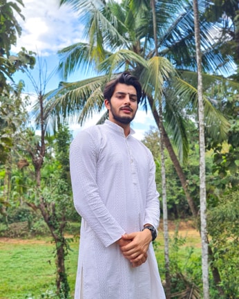 A man stands outdoors surrounded by lush greenery and tall palm trees. He is wearing a long, white, traditional South Asian attire with intricate patterns. The sky is partly cloudy, adding a soft backdrop to the vibrant green scenery.