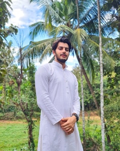 A man stands outdoors surrounded by lush greenery and tall palm trees. He is wearing a long, white, traditional South Asian attire with intricate patterns. The sky is partly cloudy, adding a soft backdrop to the vibrant green scenery.