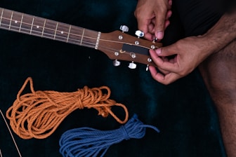 A person is adjusting the tuning pegs on a guitar headstock. Next to the guitar, there are two pieces of rope: one orange and one blue, neatly coiled on a dark surface.