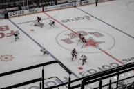 An aerial view of a hockey game in progress, featuring players in red and white jerseys on an ice rink. The surface is marked with visible lines and advertisements, including a prominent logo at center ice.