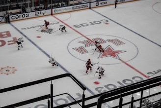 An aerial view of a hockey game in progress, featuring players in red and white jerseys on an ice rink. The surface is marked with visible lines and advertisements, including a prominent logo at center ice.