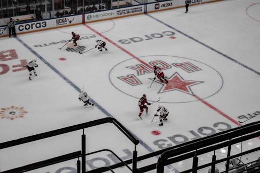 An aerial view of a hockey game in progress, featuring players in red and white jerseys on an ice rink. The surface is marked with visible lines and advertisements, including a prominent logo at center ice.