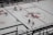 An aerial view of a hockey game in progress, featuring players in red and white jerseys on an ice rink. The surface is marked with visible lines and advertisements, including a prominent logo at center ice.