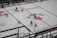 An aerial view of a hockey game in progress, featuring players in red and white jerseys on an ice rink. The surface is marked with visible lines and advertisements, including a prominent logo at center ice.