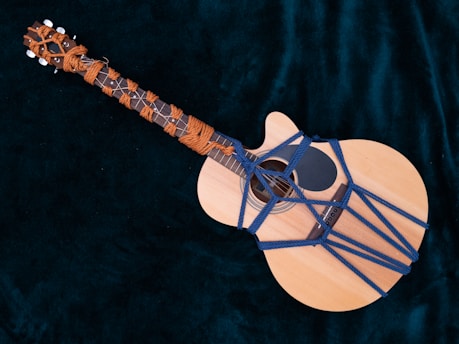 An acoustic guitar wrapped with colorful ropes in an intricate pattern, placed on a dark velvet fabric background.