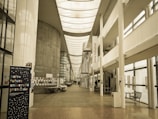 A spacious, modern university hallway featuring a high ceiling with skylights and large windows that allow natural light to illuminate the area. There are informational posters and a stand with notices along the walls. A staircase leads to upper levels, and there is seating available in the corridor. The banner in the foreground displays text in French.