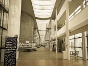 A spacious, modern university hallway featuring a high ceiling with skylights and large windows that allow natural light to illuminate the area. There are informational posters and a stand with notices along the walls. A staircase leads to upper levels, and there is seating available in the corridor. The banner in the foreground displays text in French.