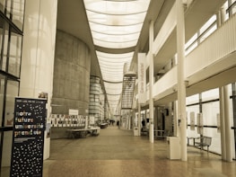 A spacious, modern university hallway featuring a high ceiling with skylights and large windows that allow natural light to illuminate the area. There are informational posters and a stand with notices along the walls. A staircase leads to upper levels, and there is seating available in the corridor. The banner in the foreground displays text in French.