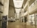 A spacious, modern university hallway featuring a high ceiling with skylights and large windows that allow natural light to illuminate the area. There are informational posters and a stand with notices along the walls. A staircase leads to upper levels, and there is seating available in the corridor. The banner in the foreground displays text in French.