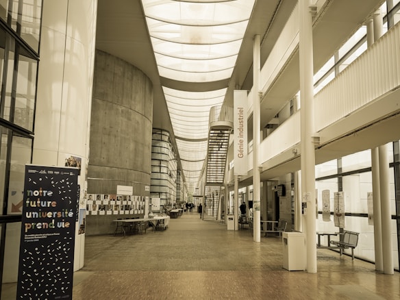 A spacious, modern university hallway featuring a high ceiling with skylights and large windows that allow natural light to illuminate the area. There are informational posters and a stand with notices along the walls. A staircase leads to upper levels, and there is seating available in the corridor. The banner in the foreground displays text in French.