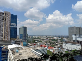 A cityscape featuring multiple high-rise buildings with varied architectural designs. The foreground includes a tall, rectangular building with a grid of windows, and a modern blue glass skyscraper displaying 'OFFICE SPACE' signage. The city stretches out into the distance, where residential and commercial structures are visible. The sky is partly cloudy, adding a dynamic element to the scene.