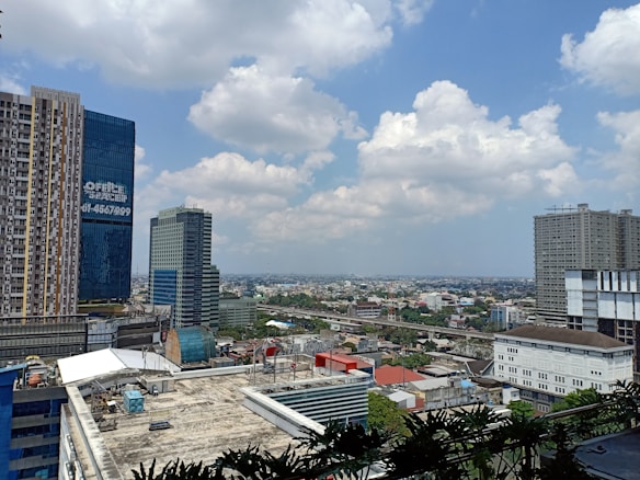 A cityscape featuring multiple high-rise buildings with varied architectural designs. The foreground includes a tall, rectangular building with a grid of windows, and a modern blue glass skyscraper displaying 'OFFICE SPACE' signage. The city stretches out into the distance, where residential and commercial structures are visible. The sky is partly cloudy, adding a dynamic element to the scene.
