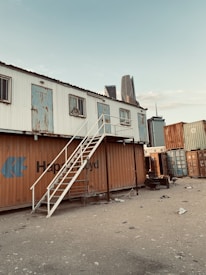 A makeshift structure constructed from shipping containers, with an external staircase leading to an upper level. The containers have varied degrees of rust and wear, and the ground is scattered with debris. Skyscrapers in the background indicate an urban setting.