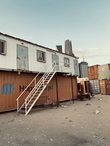 A makeshift structure constructed from shipping containers, with an external staircase leading to an upper level. The containers have varied degrees of rust and wear, and the ground is scattered with debris. Skyscrapers in the background indicate an urban setting.