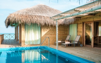 A tropical overwater bungalow constructed with wooden materials and thatched roofing sits adjacent to a pristine swimming pool. Two lounge chairs are placed on a wooden deck under a bamboo pergola, offering a relaxing spot with views of the ocean. Curtains hang in front of large glass windows, and blue skies are visible in the background.