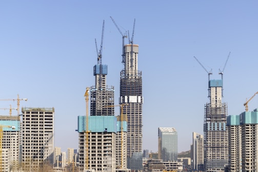 Several high-rise buildings under construction with cranes towering over them. The sky is clear and blue, providing a backdrop for the emerging skyscraper structures. The buildings exhibit different levels of completion, with scaffolding and construction platforms visible.