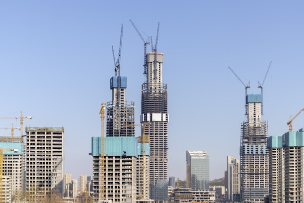 Several high-rise buildings under construction with cranes towering over them. The sky is clear and blue, providing a backdrop for the emerging skyscraper structures. The buildings exhibit different levels of completion, with scaffolding and construction platforms visible.